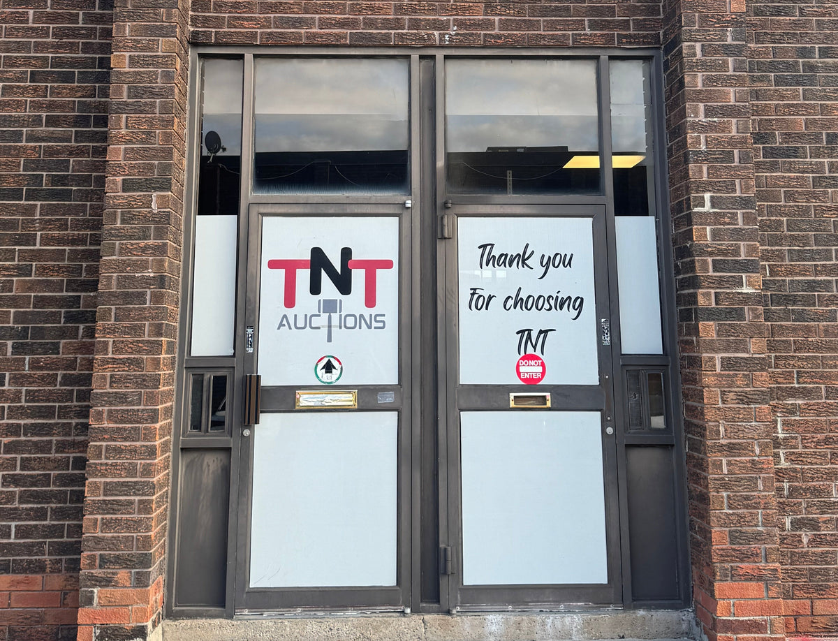 TNT Auctions storefront with double glass doors set in a red-brown brick building. One door has a white sign reading 'TNT AUCTIONS' and the other says 'Thank you for choosing TNT'.